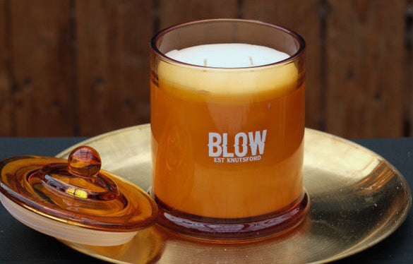 Candle in a glass jar labeled 'BLOW' on a metallic tray with a wooden background